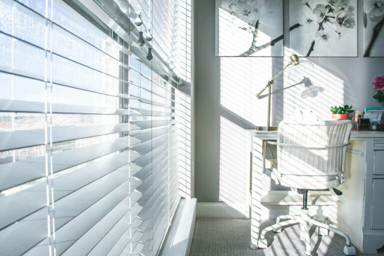 Das Bild zeigt ein Büro, in welches warme Sonnenstrahlen hereinfallen. Am Fenster sind weiße Innenjalousien von Suntec zum Sonnenschutz angebracht.