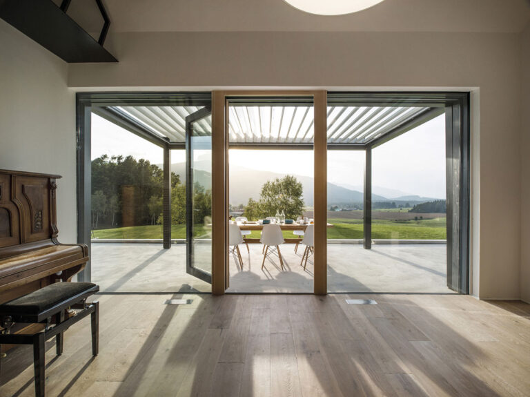 das Bild zeigt eine Aussicht aus einem Wohnraum auf eine Terrasse. Diese Terrasse im Münsterland hat ein Lamellendach von Suntec aus Coesfeld.